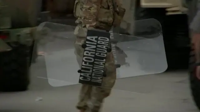 A camouflaged man carries a shield reading California National Guard