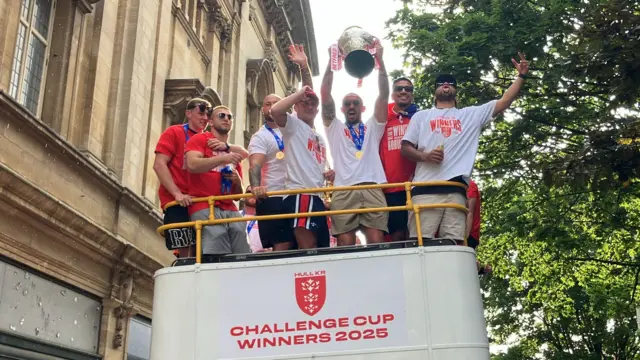 Seven Hull KR players pose on top of the open top bus as they leave the Hull City Hall. Captain Elliot Minchella, with a gold medal around his neck, is cheering as he holds a large silver trophy in the air.