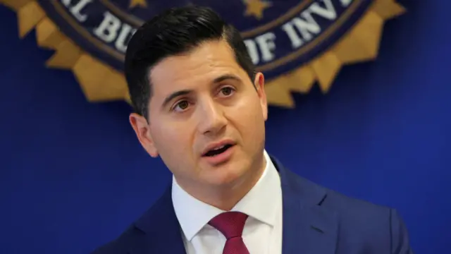 Bill Essayli, who has short black hair, wears a blue suit with a white shirt and red tie