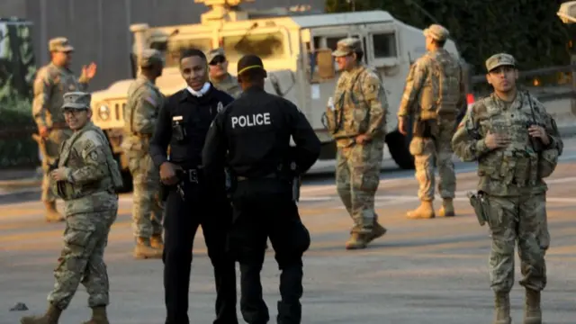 Seven members of the National Guards of California in military fatigues with two police officers stand outdoors, a military vehicle in the background