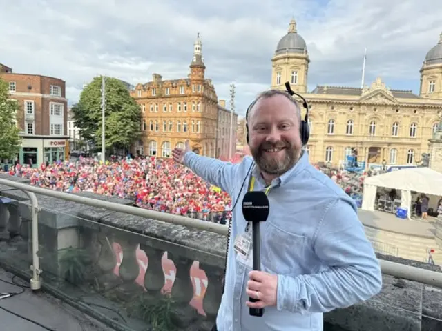 James Hoggarth is wearing a blue shirt and holding a microphone on the balcony of Hull City Hall