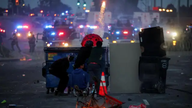 A firework is seen flying toward police vans from behind a bin where four protesters crouch. Two are seen wearing face masks, while one wears a helmet and holds up an umbrella