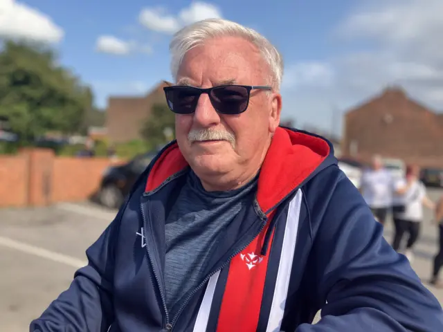 A man with grey hair and a moustache stares into the camera. He is wearing dark sunglasses and a navy blue, red and white hoodie.