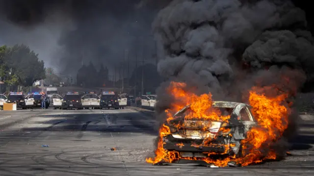 A car burns on the road near a line of police cars in LA