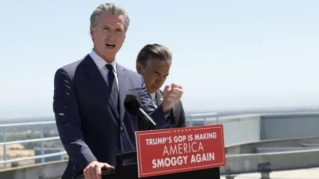 Gavin Newsom speaking into microphone, stood behind a podium with message written on it. Message says: Trump's GOP is making America smoggy again.