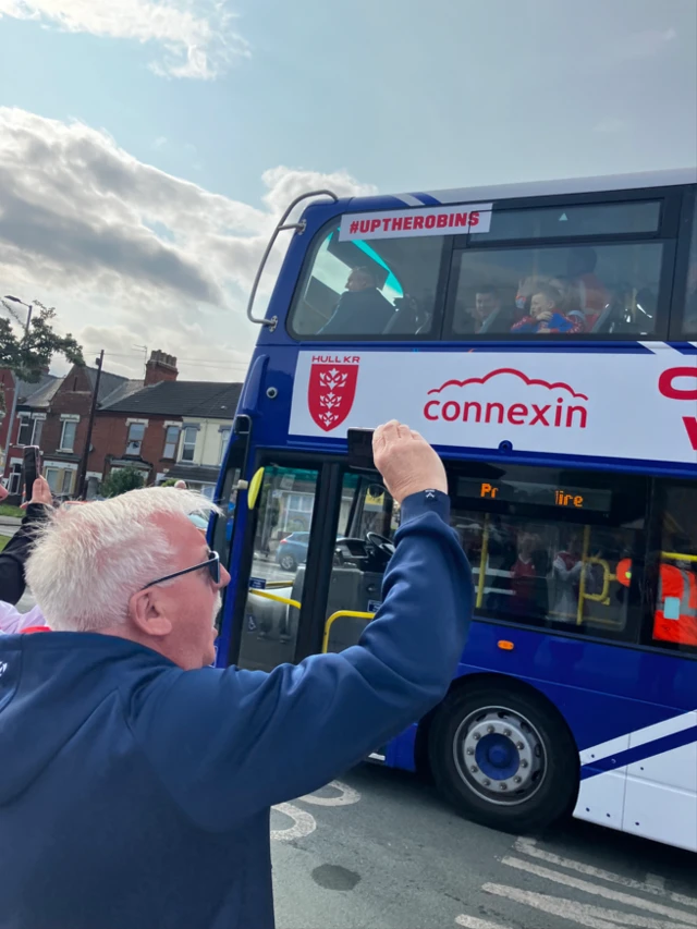 A man with grey hair raises a fist and cheers as a bus passes him