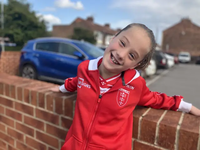 A girl with brown hair smiles as she stands against a brick wall. She is wearing a red and white hoodie with the Hull Kingston Rovers logo on it.
