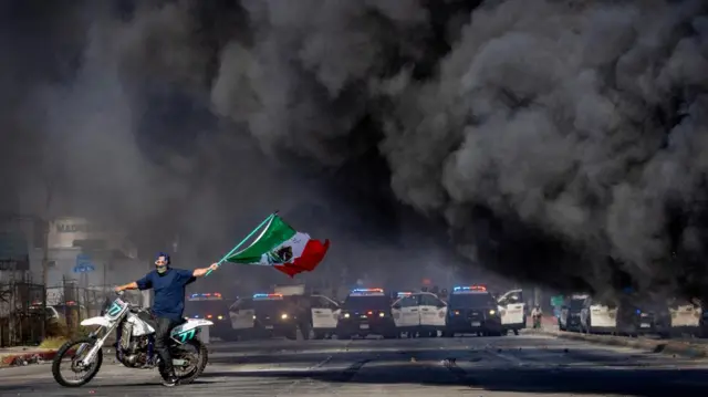 A man on a motorcycle waves a Mexican flag as smoke rises from a burning car on Atlantic Boulevard, during a standoff by protesters and law enforcement, following multiple detentions by Immigration and Customs Enforcement (ICE), in the Los Angeles County city of Compton, California, U.S., June 7, 2025