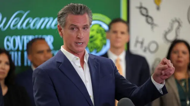California governor Gavin Newsom in dark blue suit, white shirt as he delivers speech, four people (from left to right: a brunette woman, an Afro-American man, a white man and a Latino woman) blurred behind him