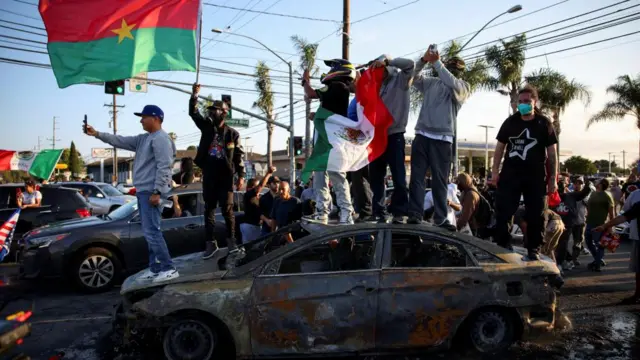 Protesters stood on a burnt out car on a street, one holds a Burkina Faso flag, another holds a Mexico flag