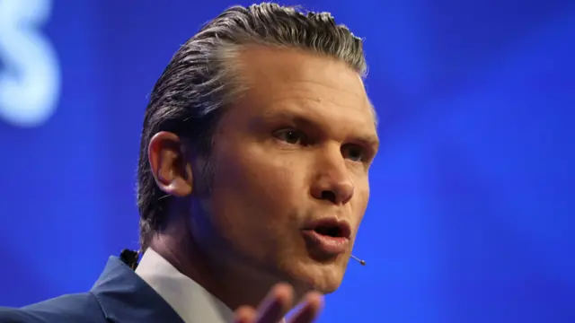 Pete Hegseth, who has slicked back grey hair, gesticulates as he speaks on stage