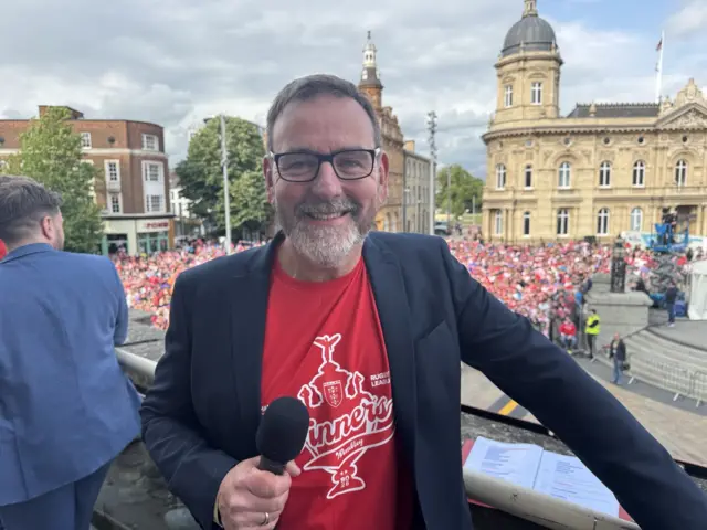 Neil holds a microphone in his hand and wears a red winners t-shirt underneath a blue jacket.  He stands on the balcony with the fans in the background