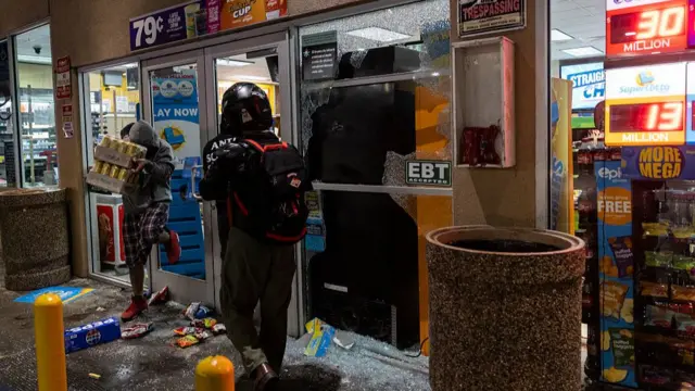 A person, whose face is covered, is seen walking out of the shattered glass door of a gas station shop, carrying two crates of drinks