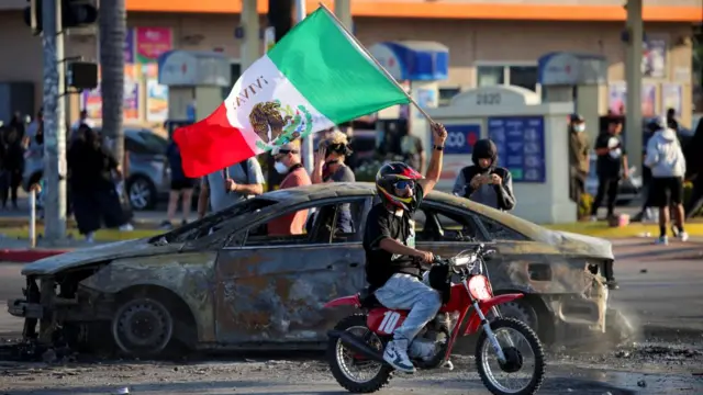 A person waves a Mexican flag while riding on a motorbike around a burned car