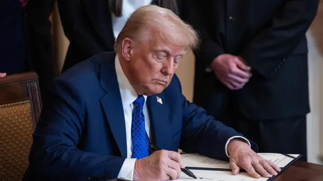 Donald Trump signs executive order on immigration. He's sitting down on a wooden chair at a wooden table wearing a dark blue suit, white shirt and royal blue tie with polka dots. A man in a black suit with his hands on top of each other in front of him stands to Trump's left, his head out of shot