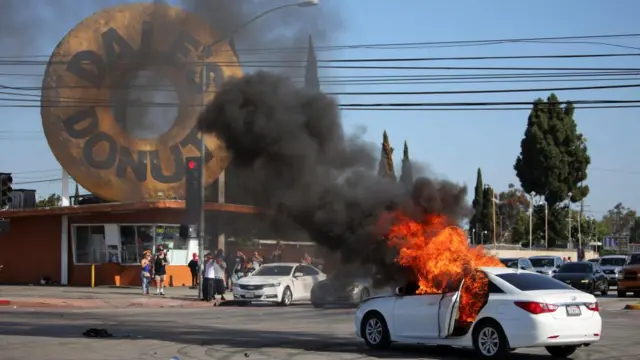 Flames are seen billowing from the open door of a white car, as onlookers watch on outside a branch of a donut store