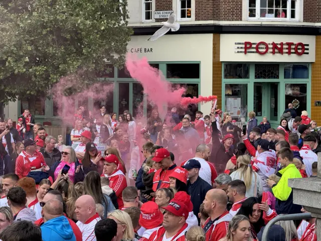A crowd of people, wearing red and white coloured clothing, stand in a public square. One man is holding a flare over his head which is emitting red coloured smoke