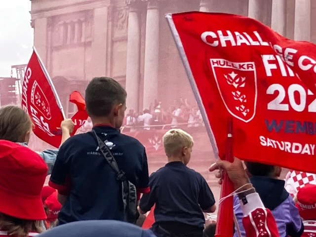 Two young fans sit on shoulders as they look towards the balcony at Hull City Hall. Through the red smoke the sillhouette of the Challenge Cup is visible in one of the players arms.