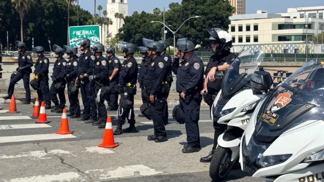 A line of a dozen or so officers with helmets stand across a road, with orange cones placed in front of them