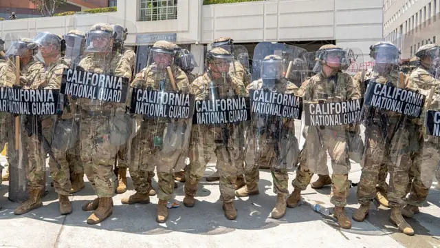 National Guard members stand in a line. they are wearing military uniforms and carry plastic shields reading 'california national guard'.