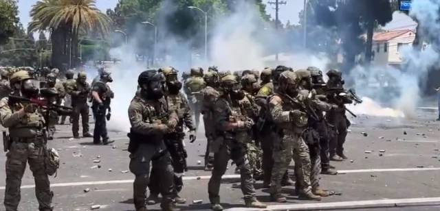 Police are seen in riot gear, with weapons, walking down a street where there is smoke. It is broad daylight.
