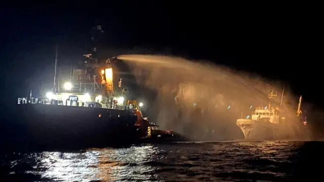 A larger boat hosing down a smaller boat in the night, illuminated by a bright orange light from the larger boat