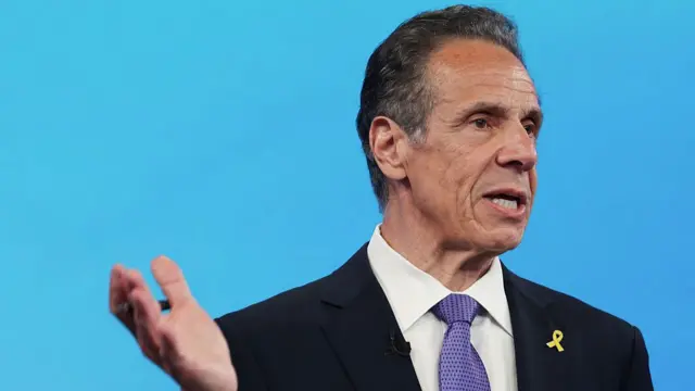 Andrew Cuomo stands in front of a blue wall wearing a black suit, white shirt and purple tie with polka dots, a yellow ribbon pin on his left lapel