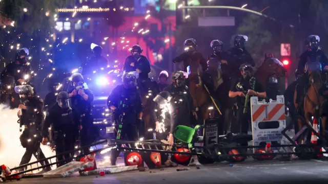 Police officers, some on horseback, are seen in LA at night, with sparks flying around them as protests continue