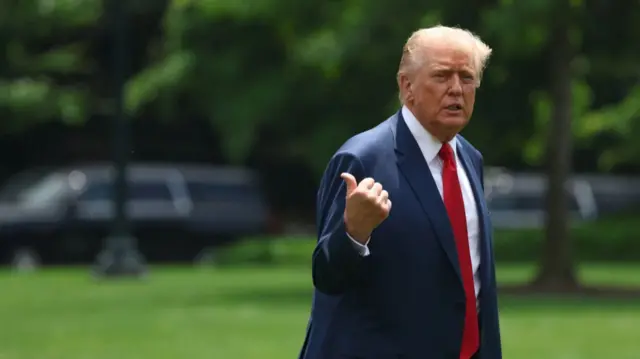 Donald Trump in dark blue suit, white shirt and red tie walks on the White House back lawn as he points to his back with his right hand, thumb extended