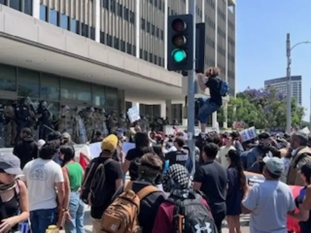 A crowd gathers outside an LA building. One man in a black shirt and backpack climbs up a traffic light