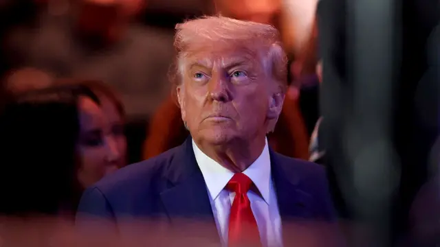 Close up of Donald Trump sitting down wearing a dark blue suit, white shirt and red tie as he watches a wrestling match inside an arena
