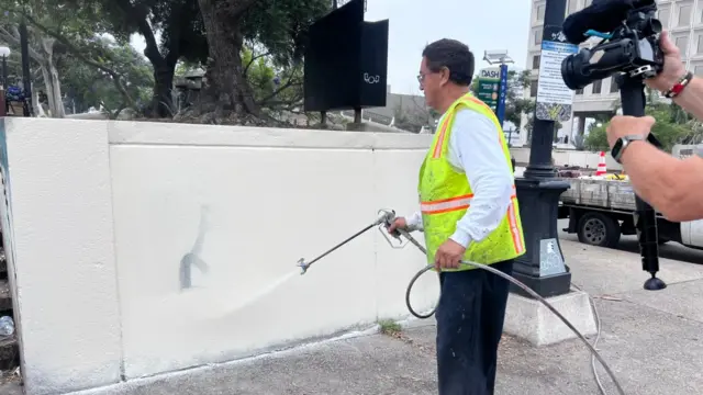 A man in a yellow Hi-Vi jacket cleans up a graffiti covered wall with a paint gun, a man holding a video camera recording him