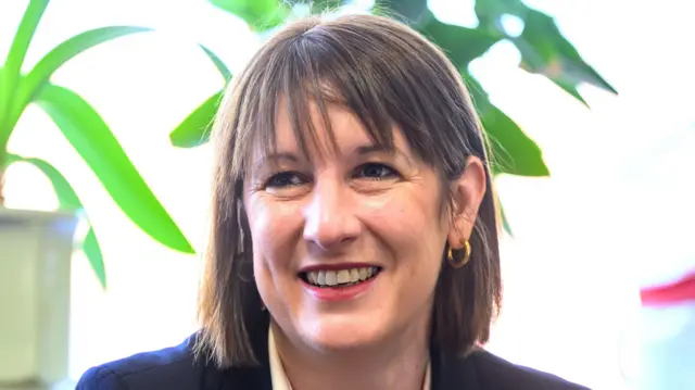 Rachel Reeves smiles as she looks away from the camera. She has brown hair that is in a bob style and she wears gold hoop earring and a dark coloured blazer with a white shirt.