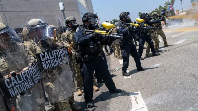 Two National Guard members in full military gear hold up plastic shields marked California National Guard. Next to them are members of the LAPD in black uniform holding up rubber bullet guns