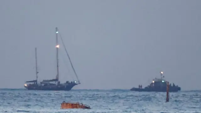 A boat that resembles the images seen earlier of the Madleen seen at sea. There appears to be a tow boat ahead. The lighting is low and the picture is taken from far away