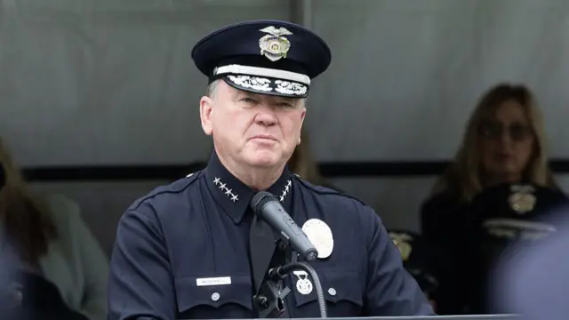 LAPD chief Jim McDonnell in black uniform shirt, four golden stars on each side of his shirt collar. He's standing at a lectern delivering a speech, other officers and people sitting under a white tent behind him (blurred)