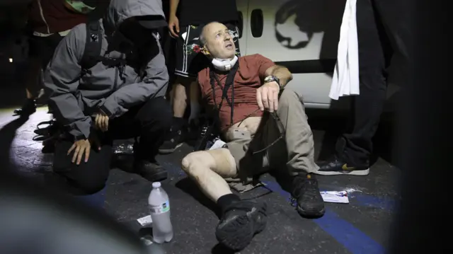 Stern, wearing a red t-shirt and cargo trousers, is treated by medics in grey jackets. it is night time and his illuminated by a camera flash.