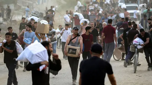 A wide shot of people, mostly men, walking along a road, carrying boxes and bags.