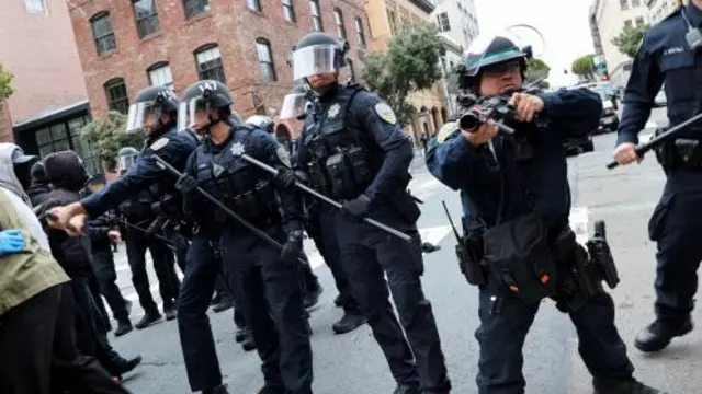Police officers stand guard during a protest against immigration sweeps at the ICE building in San Francisco. They wear an all black uniform, with helmets, holding batons and rubber bullet guns.