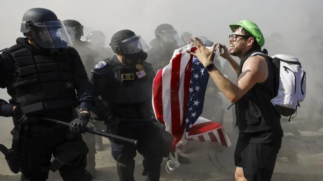 Two masked police officers square off next to a protester holding an American flag.