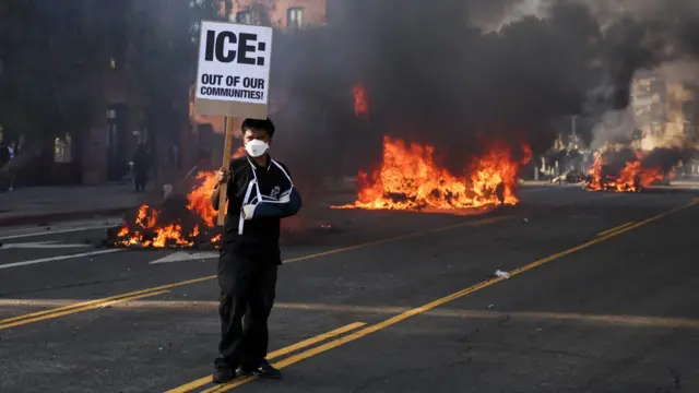 A protester holding a sign reading "ICE: Out of our communities" stands before burning debris on a Los Angeles street.