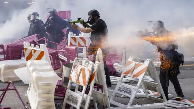Police fire less-lethal rounds at protesters from behind a makeshift barricade. Police officers in riot gear can be seen pointing guns towards protesters, while streaks of sparks are shown in front of them.
