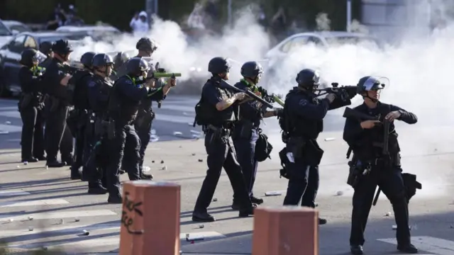 A row of officers, dressed in black with helmets, aiming rubber bullet guns, on the street with smoke.