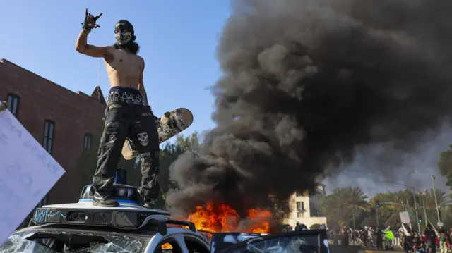 A shirtless man holding a skateboard stands on top of a damanged vehicle. Behind him, plumes of black smoke rise from another burning vehicle.