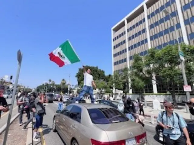 A man carrying a Mexican flag stands on top of a tan car as photographers and protestors stand around him