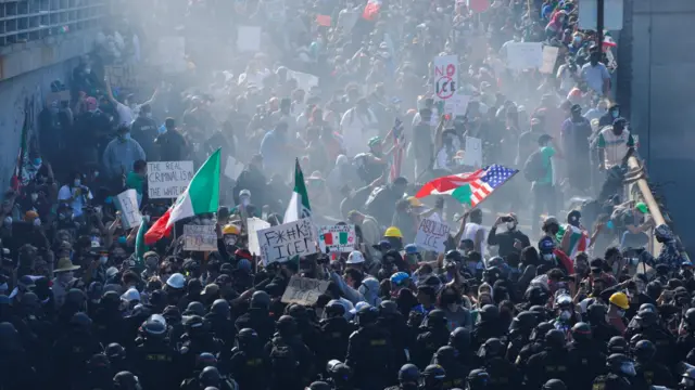 A crowd of security personnel enforce a barrier in front of protesters, shrouded in smoke. Some of the protesters are holding Mexican and US flags.