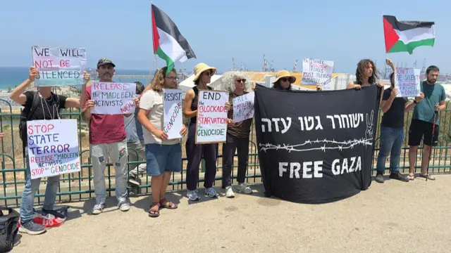 A small group of deminstratoers holding banners. Two hold up a big black flag with Free Gaza written underneath Hebrew text and an illustration of barbed wire.