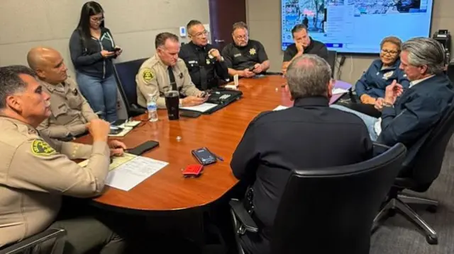 Officials sit around a board room table. they are taking notes on notepads and mobile phones. a screen shows information about the protests on the wall.