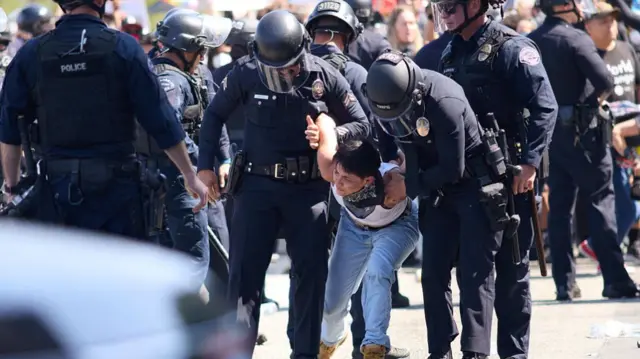 A protester wearing denim and a white vest is detained by three police officers wearing riot helmets and dressed in black.