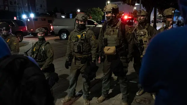 ICE agents form a human barrier on the street. They're wearing green military wear with hard helmets and coverings over their mouths and noses